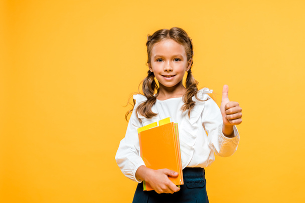 happy kid holding books and showing thumb up isolated on orange  - 写真・画像