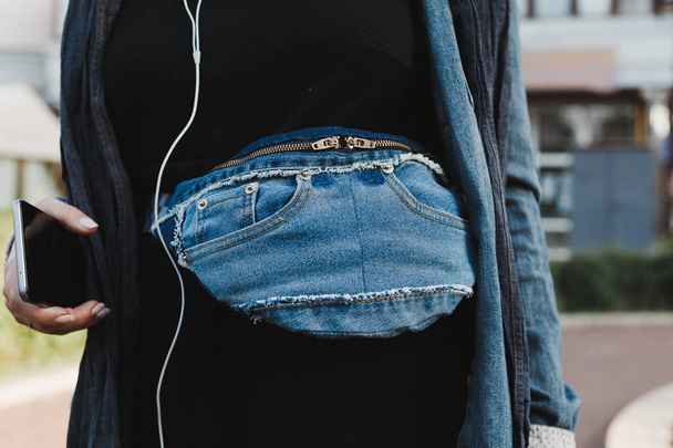 Modny niebieski stylowy pasek torba denim sprzęgła na młodej kobiety z telefonem w ręku i drutu słuchawek. Miejski styl uliczny - Zdjęcie, obraz