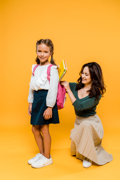 attractive mother putting books in backpack of schoolgirl on orange  - Foto, Bild