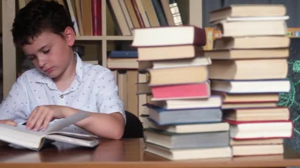 boy with dark hair, light shirt sits at table and reads book. Misses. Leafing through pages relying hand on cheek. lot of books in stack of books. Panoramic shooting on background of the bookshelf. - Footage, Video