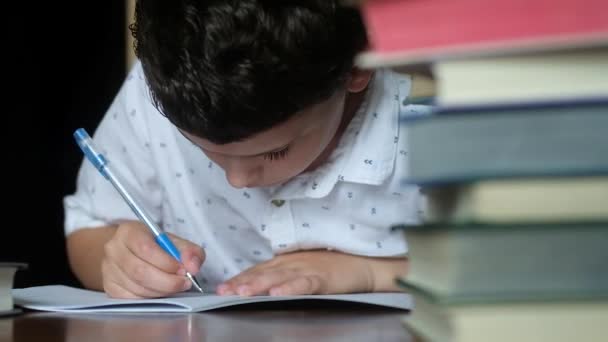 lähikuva pojasta, joka istuu puisessa pöydässä valkoisessa paidassa. Hän kirjoittaa kynällä, jossa on sininen hattu muistikirjassa. Ratkaisee luokkahuoneen ongelmat. Paljon koulukirja pino kirjoja
. - Materiaali, video