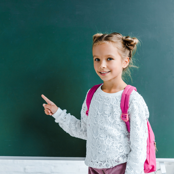 happy kid smiling while pointing with finger at green chalkboard  - Foto, Bild