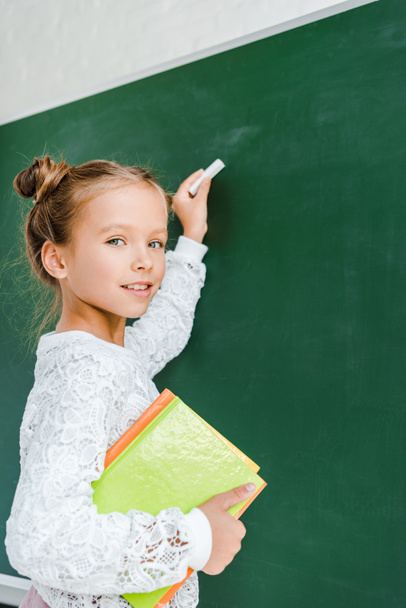 happy schoolkid holding chalk and books near green chalkboard  - Фото, изображение