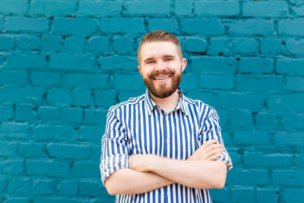 Πορτρέτο ενός νεαρού θετικού κομψού χαμογελαστή άντρα με μουστάκι και γενειάδα στο φόντο ενός μπλε τοίχου με τούβλα. Η ιδέα των διακοπών στην πόλη. - Φωτογραφία, εικόνα