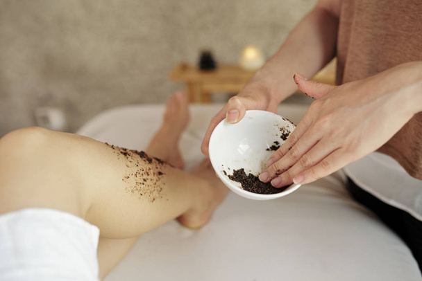Imagem de close-up de cosmetologista aplicando esfoliação de damasco natural em pernas de jovem relaxante no salão de spa
 - Foto, Imagem
