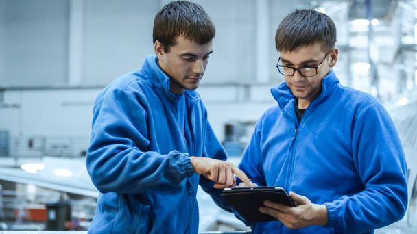Two aircraft maintenance mechanics have a conversation while using a tablet in a plane hangar. - Photo, Image