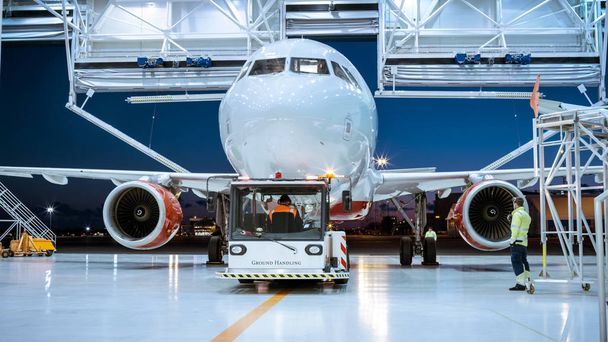 Aircraft Maintenance Hangar Where New Airplane is Toed by a Pushback Tractor/ Tug onto Landing Strip. Crew of Mechanics, Engineers and Drivers Works Busily. - Photo, Image