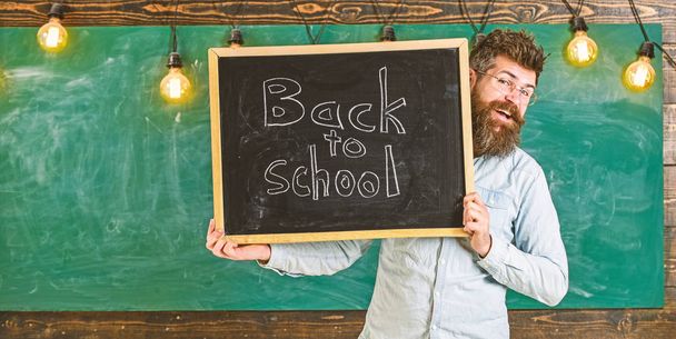Start of school year concept. Teacher on cheerful face holds blackboard with title back to school. Man with beard welcomes students, chalkboard on background. Teacher peeking out of chalkboard - Фото, изображение