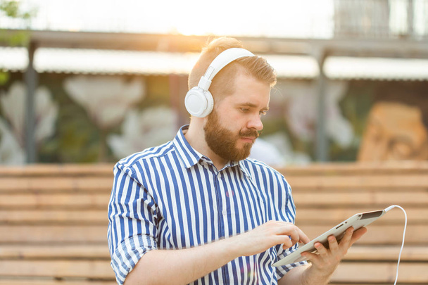Serious stylish young male student listens to lectures using tablet headphones and high-speed wireless internet while sitting on a wooden park bench on a summer day. Online learning concept. - Foto, afbeelding