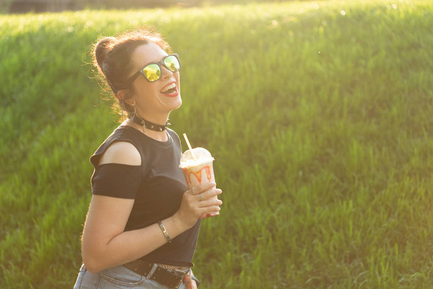 Pozytywna, wesoła młoda kobieta w stylowych punkowych ubraniach i okularach trzyma koktajl w rękach podczas spaceru w parku w ciepłym letnim wieczorem. Koncepcja relaksu i zabawy w mieście. - Zdjęcie, obraz