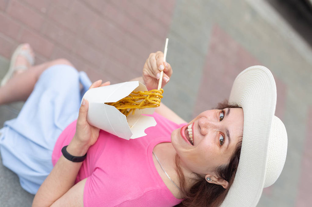 Close-up of young woman in a hat eats Chinese noodles while walking in the park on a warm summer day. Takeaway food concept. - Photo, Image