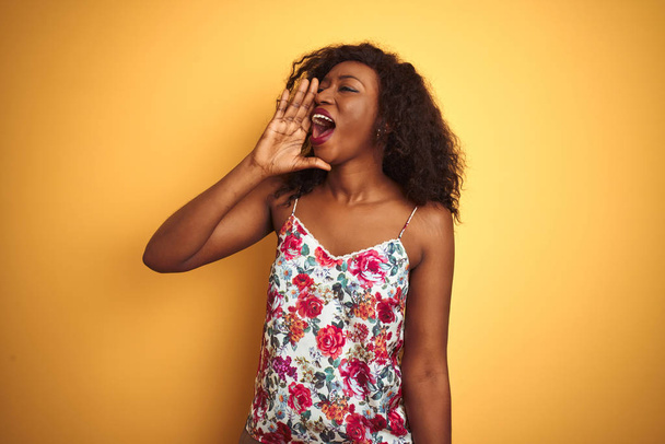 Femme afro-américaine portant un t-shirt floral d'été sur fond jaune isolé criant et criant haut et fort à côté avec la main sur la bouche. Concept de communication
. - Photo, image