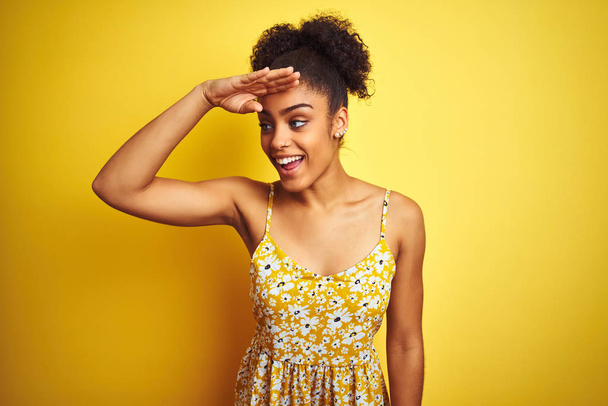 Afro-amerikai nő visel alkalmi virágos ruha állt át elszigetelt sárga háttér nagyon boldog, és mosolyogva keres messze a kezét a feje fölött. Keresés a koncepcióban. - Fotó, kép