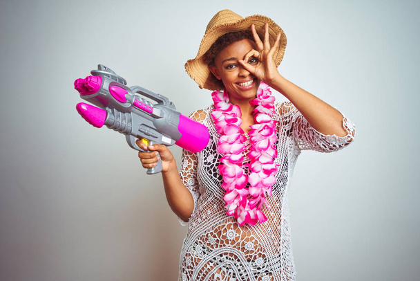 Молодая африканская американка с афроволосами в цветочном гавайском леи и водяным пистолетом со счастливым лицом, улыбающаяся, делает хорошо знак с рукой на глазу, глядя сквозь пальцы
 - Фото, изображение