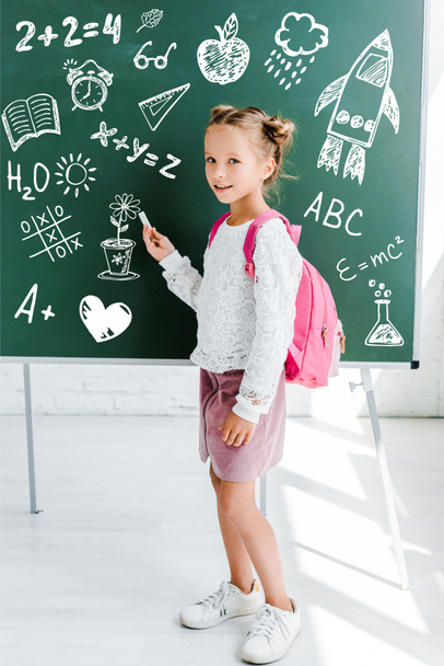 heureux enfant debout avec sac à dos et tenant craie près de dessin sur tableau vert
  - Photo, image