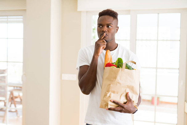 Afro-amerikai férfi gazdaság papírzacskót tele friss élelmiszereket komoly arca gondolkodás kérdés, nagyon zavaros ötlet - Fotó, kép