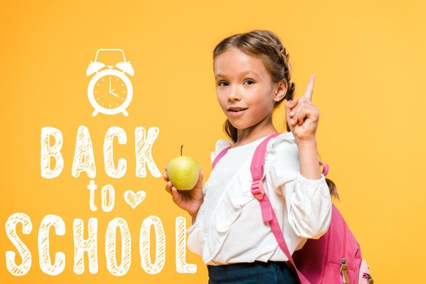 adorable schoolkid holding apple and pointing with finger near back to school lettering on orange  - Fotografie, Obrázek