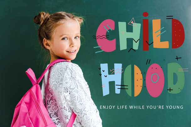 happy schoolkid smiling while standing with backpack near childhood lettering on green  - Foto, Imagem