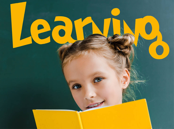 happy kid smiling while holding yellow book near learning lettering on green  - Photo, image