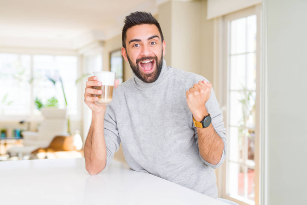 Knappe Spaanse man drinken van een kopje koffie schreeuwen trots en het vieren van de overwinning en succes zeer opgewonden, juichen emotie - Foto, afbeelding