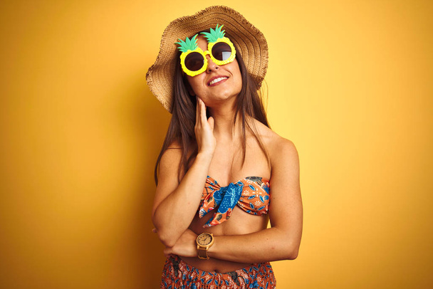 Woman on vacation wearing bikini and pineapple sunglasses over isolated yellow background with hand on chin thinking about question, pensive expression. Smiling with thoughtful face. Doubt concept. - Foto, Imagem