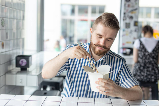 Un jeune homme élégant et positif qui mange des nouilles chinoises dans un café pendant une pause au travail. Le concept de repos et de collation saine
. - Photo, image