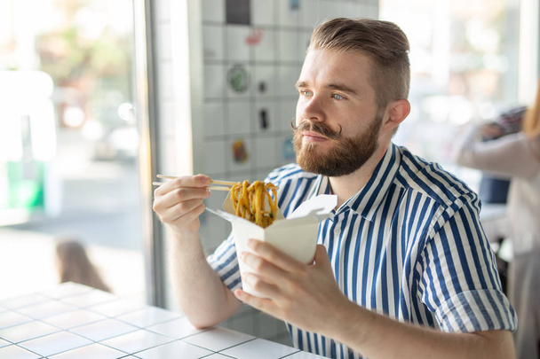 Позитивный молодой стильный парень ест китайскую лапшу в кафе во время перерыва на работе. Концепция отдыха и здоровых закусок
. - Фото, изображение