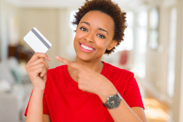 Joven mujer afroamericana sosteniendo tarjeta de crédito muy feliz señalando con la mano y el dedo
 - Foto, Imagen