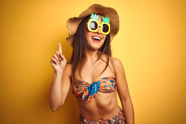 Donna in vacanza indossando bikini e occhiali da sole ananas su sfondo giallo isolato puntando il dito verso l'alto con l'idea di successo. Uscito e felice. Numero uno.
. - Foto, immagini