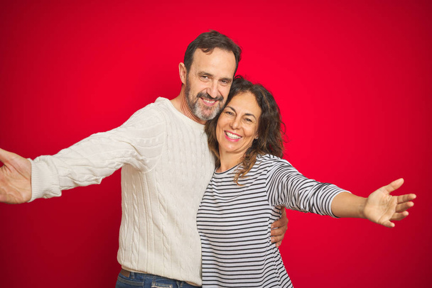 Красива пара середнього віку носить зимовий светр над ізольованим червоним тлом, дивлячись на камеру, посміхаючись з відкритими руками для обіймів. Веселий вираз, що охоплює щастя
. - Фото, зображення