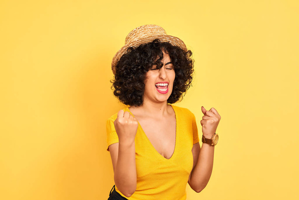 Jeune femme arabe aux cheveux bouclés portant t-shirt et chapeau sur fond jaune isolé très heureux et excité de faire geste gagnant avec les bras levés, souriant et criant pour le succès. Concept de célébration
. - Photo, image