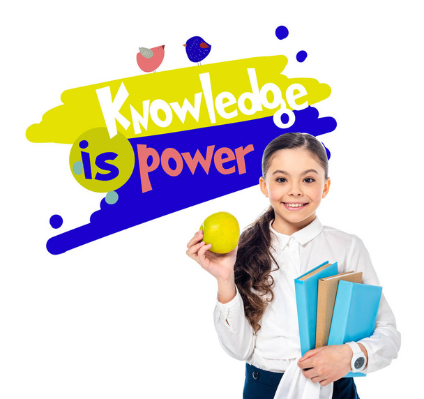 smiling schoolgirl holding apple and books while looking at camera near knowledge is power letters on white  - Foto, immagini
