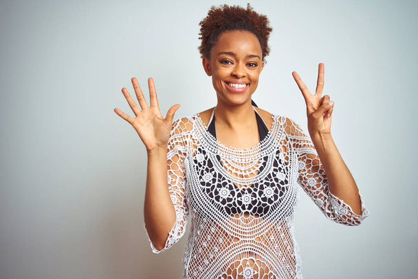 Joven mujer afroamericana con cabello afro vistiendo un bikini sobre fondo blanco aislado mostrando y señalando con los dedos número siete mientras sonríe confiada y feliz
. - Foto, imagen