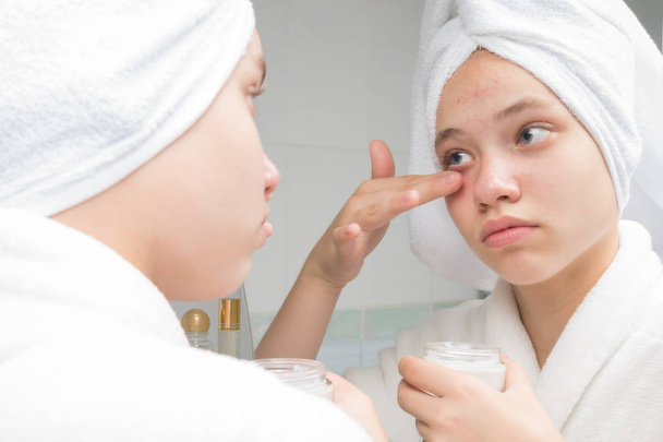 girl stands in front of a mirror in the bathroom and spreads face cream - Фото, изображение