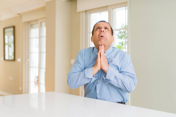 Mann mittleren Alters sitzt zu Hause und bettelt und betet mit Händen und hoffnungsvollem Gesichtsausdruck sehr emotional und besorgt. Bitte um Vergebung. Religionskonzept. - Foto, Bild