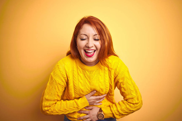 Mooie redhead vrouw dragen winter trui staande over geïsoleerde gele achtergrond glimlachend en lachen hard uit luid omdat grappige gekke grap met handen op lichaam. - Foto, afbeelding