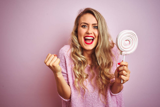 Nuori kaunis nainen syö makeaa karkkia vaaleanpunainen eristetty tausta huutaa ylpeä ja juhlii voittoa ja menestystä hyvin innoissaan, hurraavat tunteet
 - Valokuva, kuva