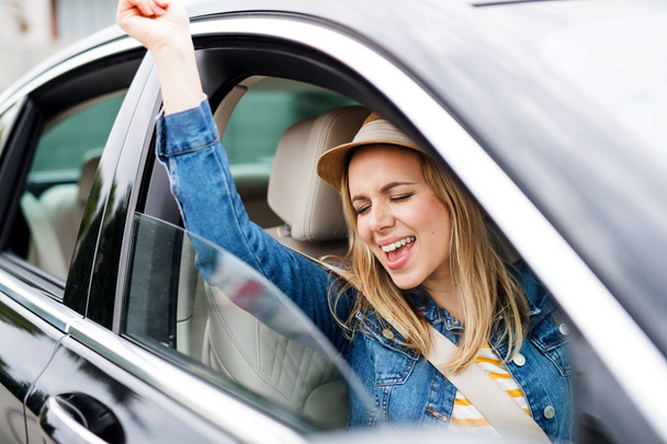 Young woman with hat sitting in car, listening to music and dancing. - 写真・画像