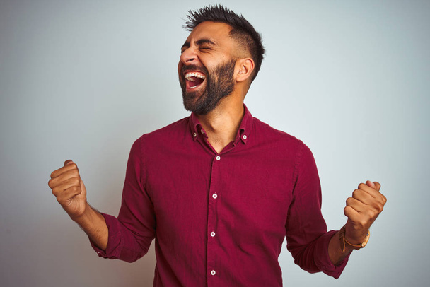 Young indian man wearing red elegant shirt standing over isolated grey background celebrating surprised and amazed for success with arms raised and eyes closed. Winner concept. - Foto, Imagem