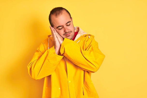 Fiatal férfi visel eső kabát állt át elszigetelt sárga háttér alszik fáradt álmodik, és pózol kézzel együtt mosolyogva csukott szemmel. - Fotó, kép