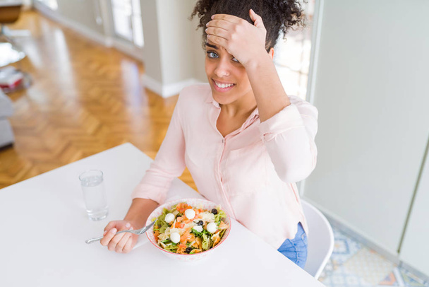 Jonge Afro-Amerikaanse vrouw die gezonde Pasta Salade eet, gestrest met de hand op het hoofd, geschokt door schaamte en verrassings gezicht, boos en gefrustreerd. Angst en boos voor fout. - Foto, afbeelding