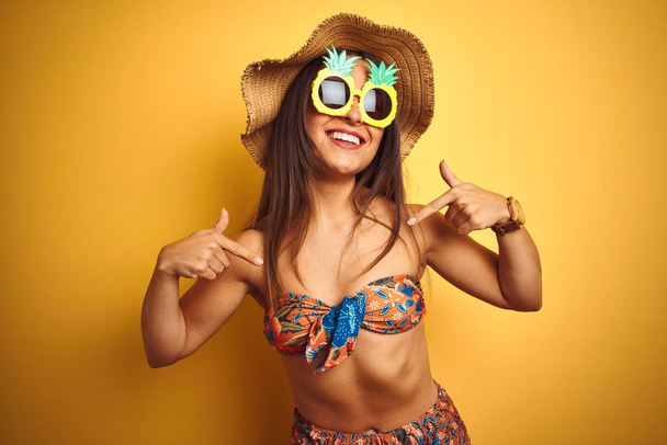 Bikini ve ananas güneş gözlüğü takan tatilde olan kadın izole sarı arka plan üzerinde yüzünde gülümseme ile emin görünümlü, parmakları gururlu ve mutlu kendini işaret. - Fotoğraf, Görsel