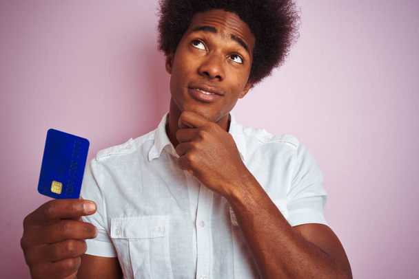 Afro-amerikanischer Kunde Mann mit Kreditkarte über isoliertem rosa Hintergrund stehend ernstes Gesicht über Frage nachdenkend, sehr verwirrte Idee - Foto, Bild