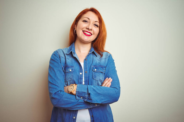Jonge mooie redhead vrouw dragen denim shirt staande over witte geïsoleerde achtergrond gelukkig gezicht glimlachend met gekruiste armen kijken naar de camera. Positieve persoon. - Foto, afbeelding