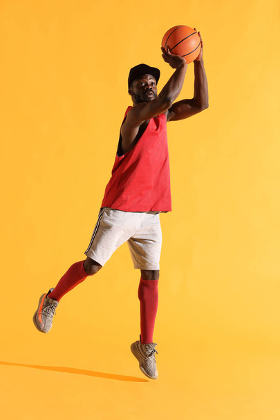Full length portrait of black man in red shirt, black cap and grey shorts playing basketball. Studio, yellow background - Foto, Imagem