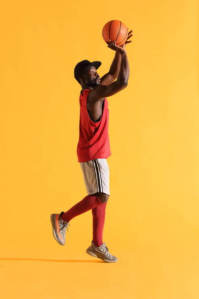 Full length portrait of black man in red shirt, black cap and grey shorts playing basketball. Studio, yellow background - Фото, зображення