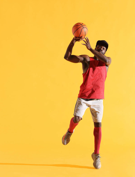 Full length portrait of black man in red shirt, black cap and grey shorts playing basketball. Studio, yellow background - Valokuva, kuva