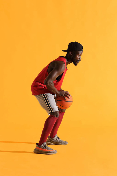 Full length portrait of black man in red shirt, black cap and grey shorts playing basketball. Studio, yellow background - Foto, immagini