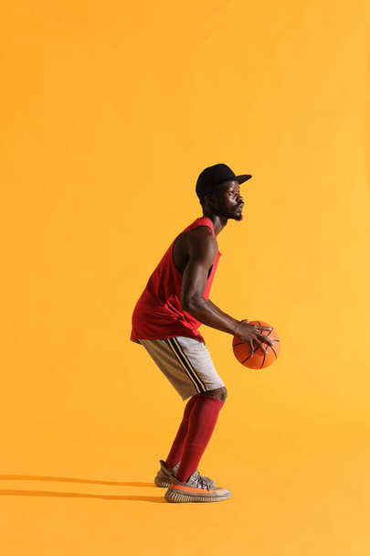 Full length portrait of black man in red shirt, black cap and grey shorts playing basketball. Studio, yellow background - Foto, imagen