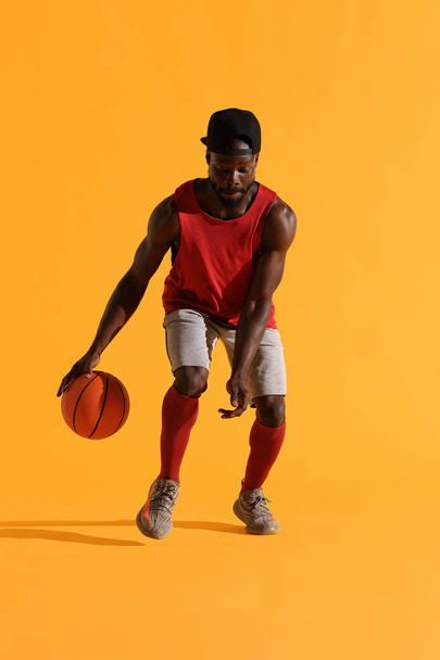 Full length portrait of black man in red shirt, black cap and grey shorts playing basketball. Studio, yellow background - Φωτογραφία, εικόνα